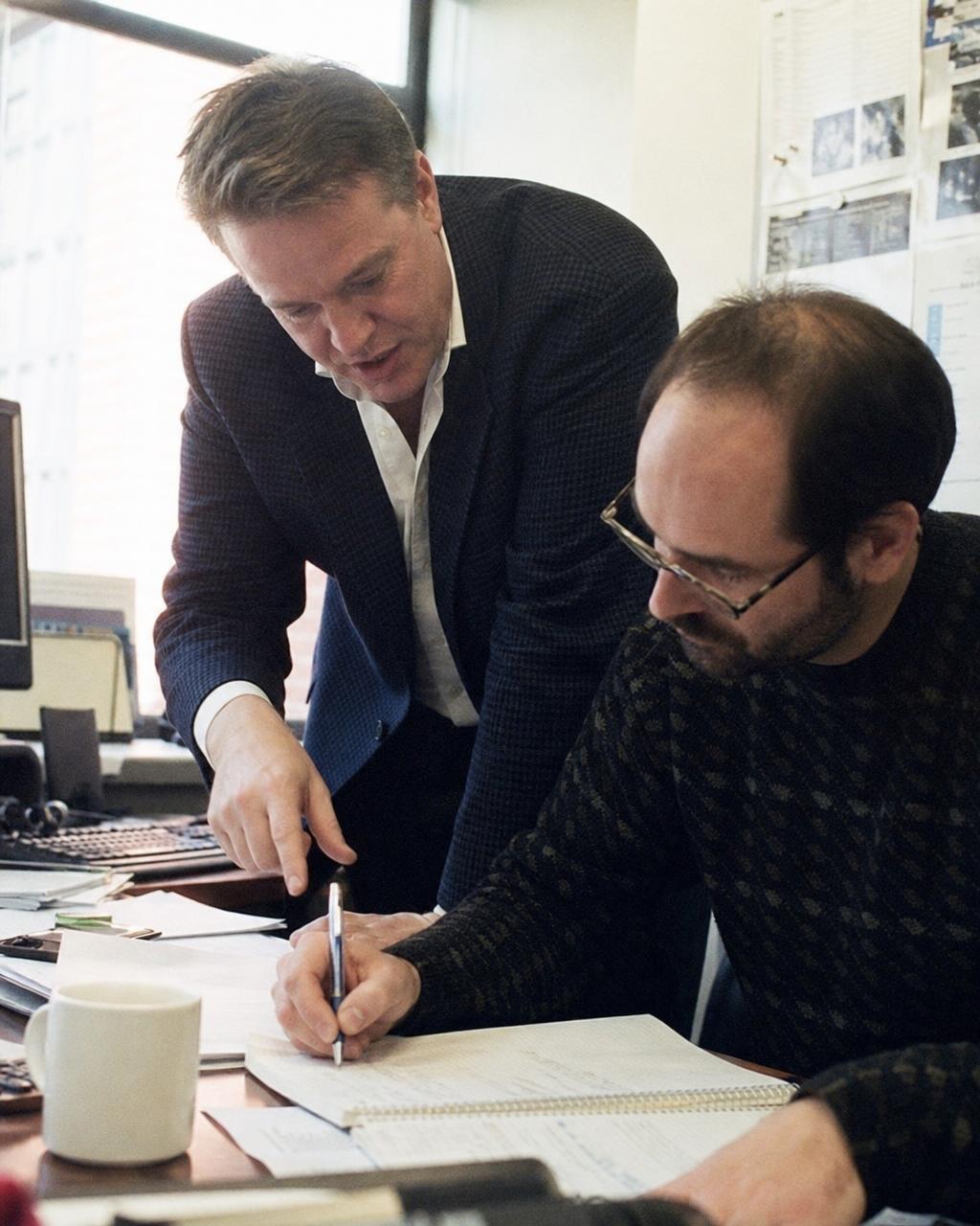 A manager leaning over a desk pointing at a notepad while a seated teammate takes notes